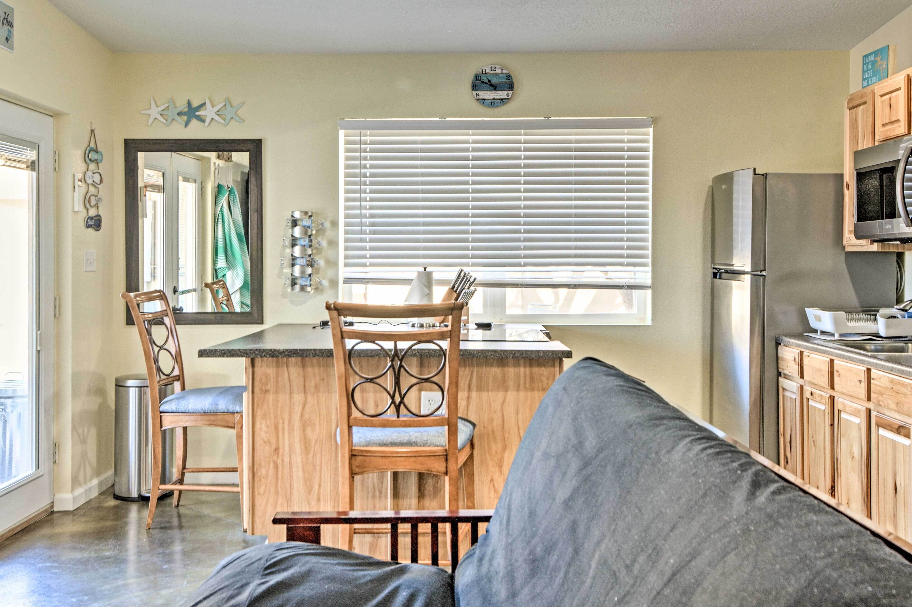 A compact kitchen with a refrigerator, microwave, and wooden cabinets. There is a window with blinds, a sink, and a chair with a unique back design.