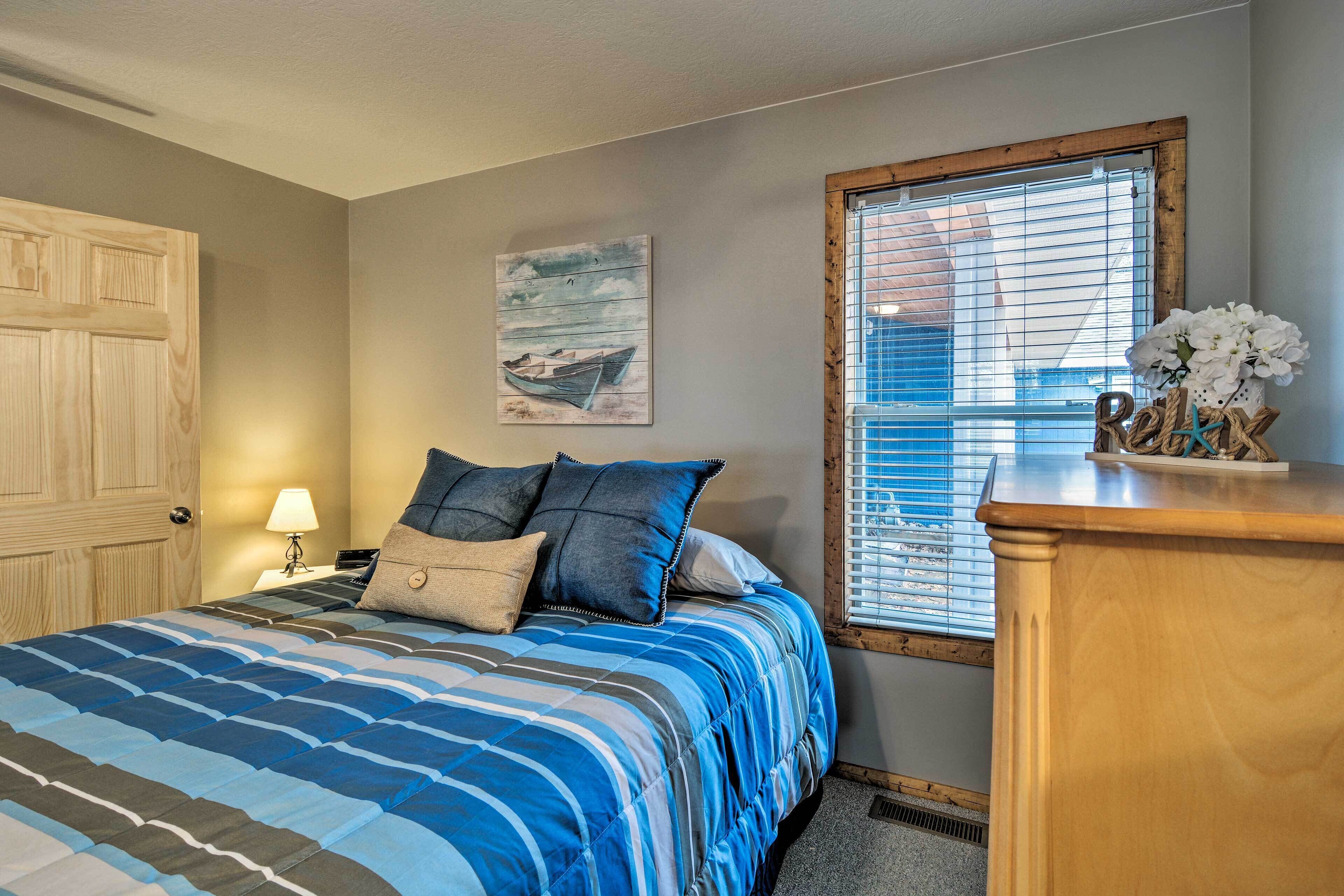 A bedroom with a bed, a nightstand, a wooden dresser, a window with blinds, and a wall-mounted painting.