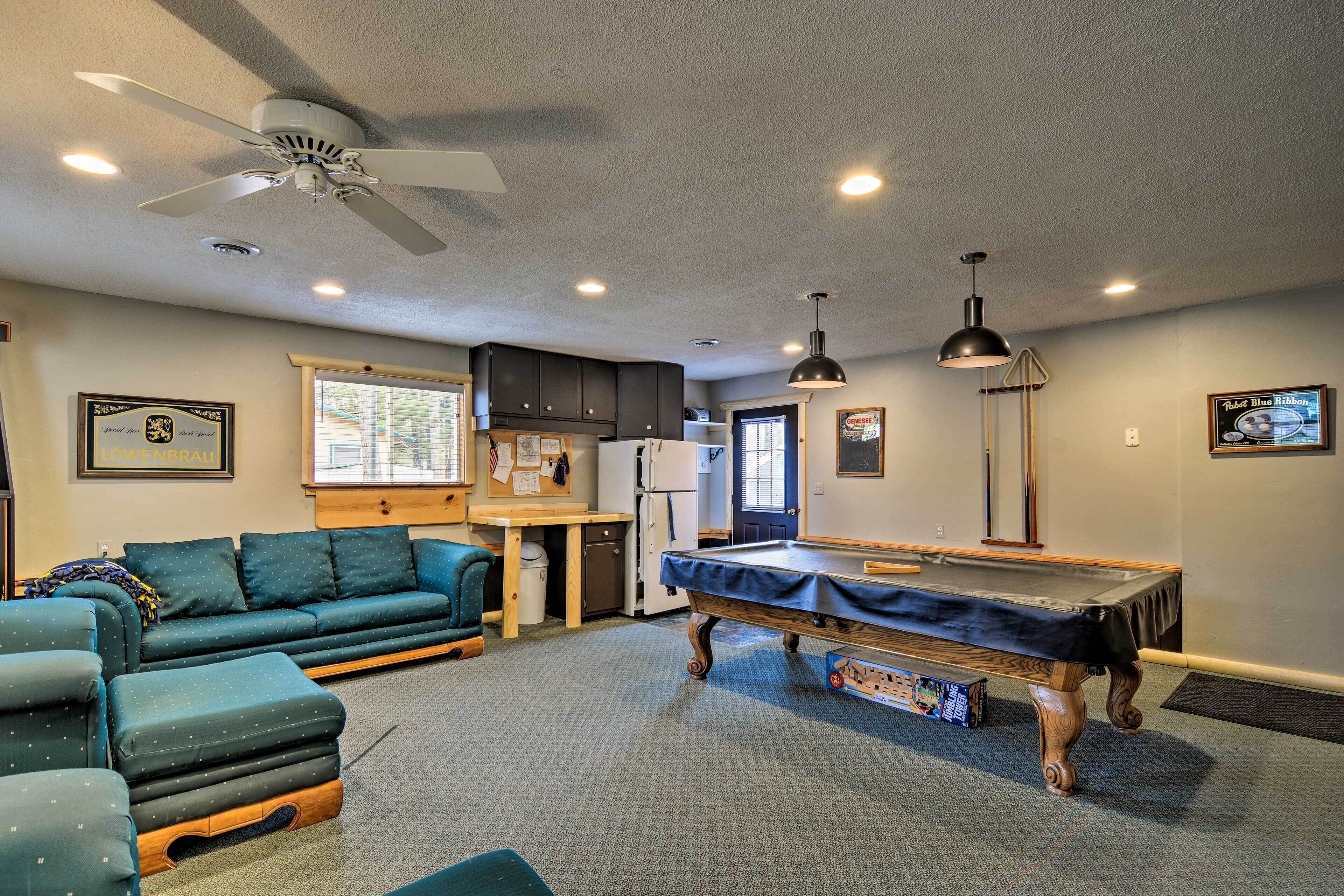 A room with a pool table, a sofa, a ceiling fan, and a kitchen area.