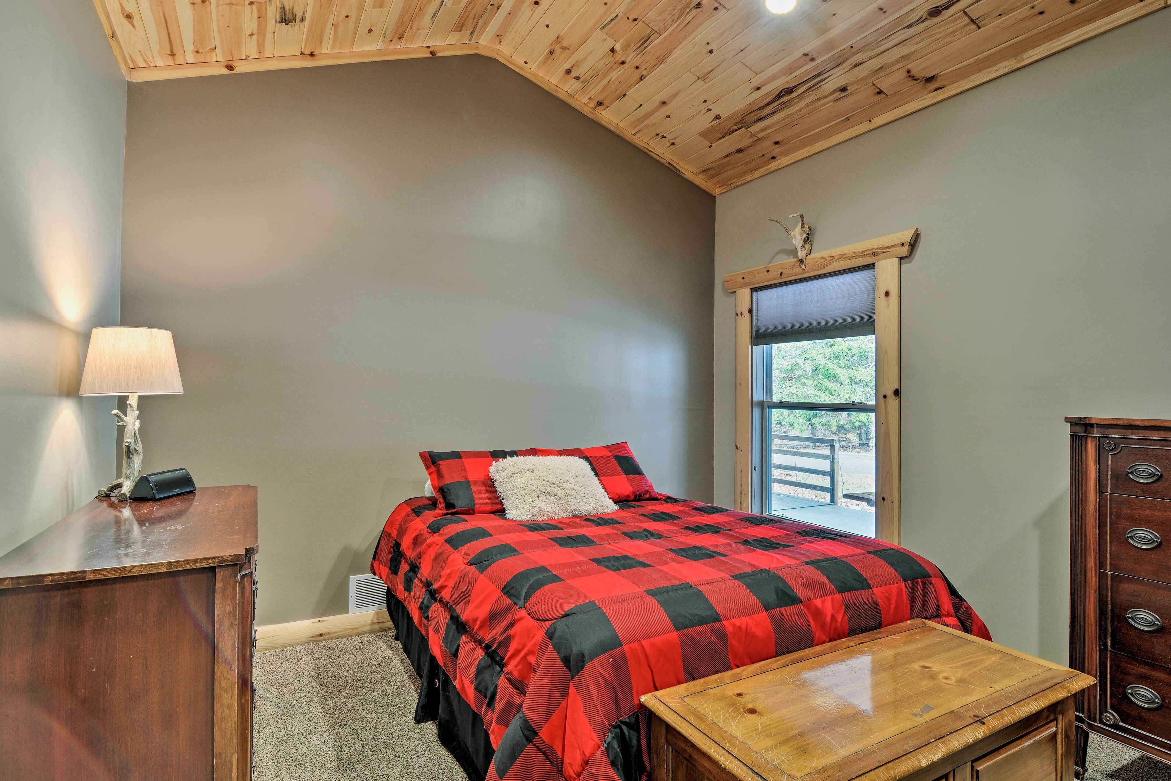 A bedroom with a bed, a dresser, a nightstand, a window, and a ceiling with wooden beams.