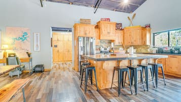 Eine geräumige Küche mit Holzschränken, einer großen Kochinsel mit Barhocker, einem Kühlschrank und einem Fenster mit Blick auf Grünflächen.