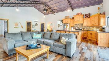 Ein geräumiges Wohnzimmer mit einem grauen Ecksofa, einem Holztisch und einer Küchenzeile mit Holdekor und einer Steinrückwand.
