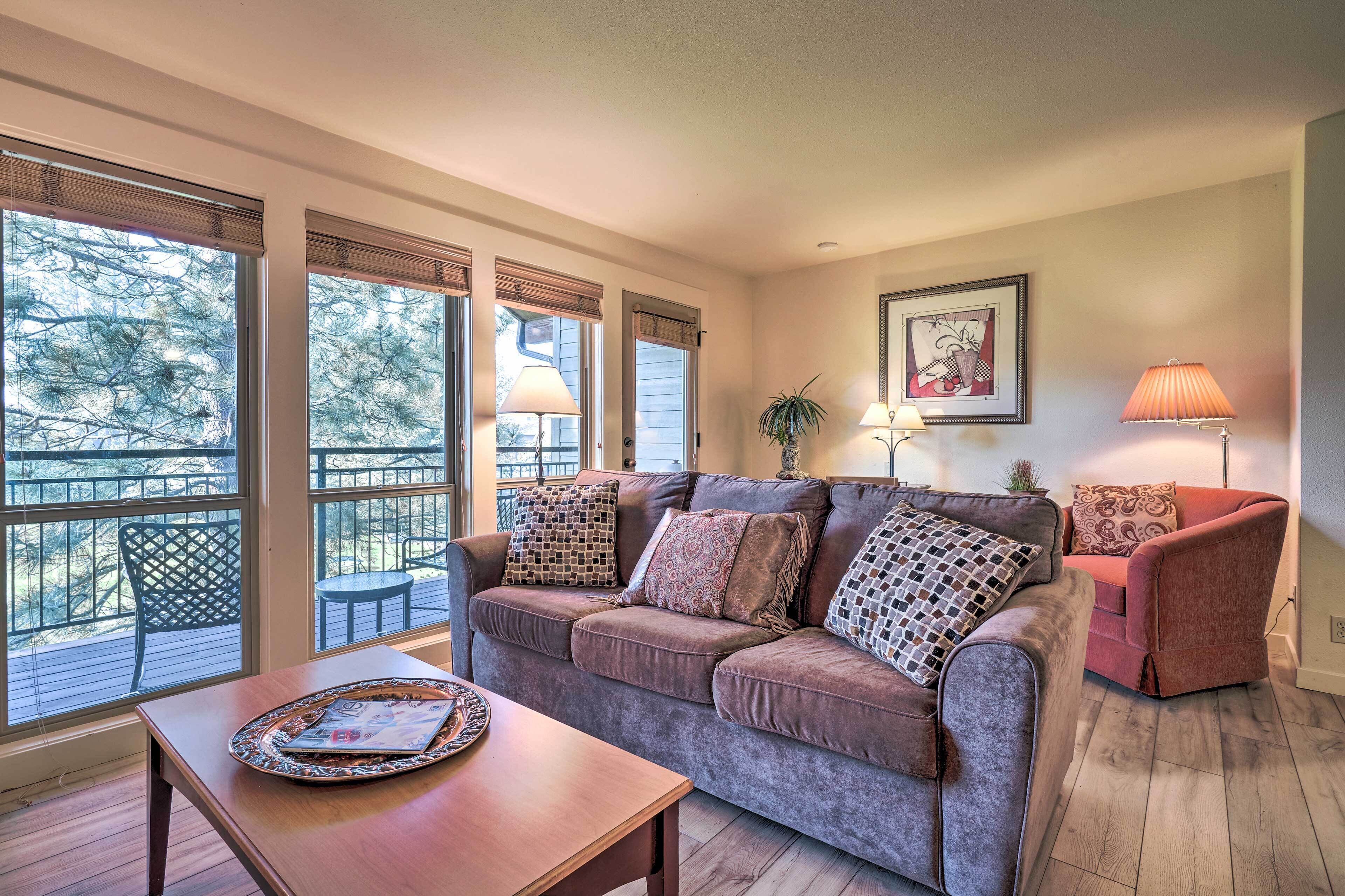 A cozy living room with a sofa, a coffee table, and two lamps. Large windows with blinds provide a view of the outdoors.