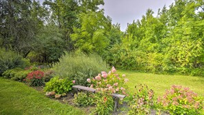 A garden with a wooden bench, various flowering plants, and dense trees in the background. - Auburn Vacation Rental Home Near Owasco Lake! (Auburn)