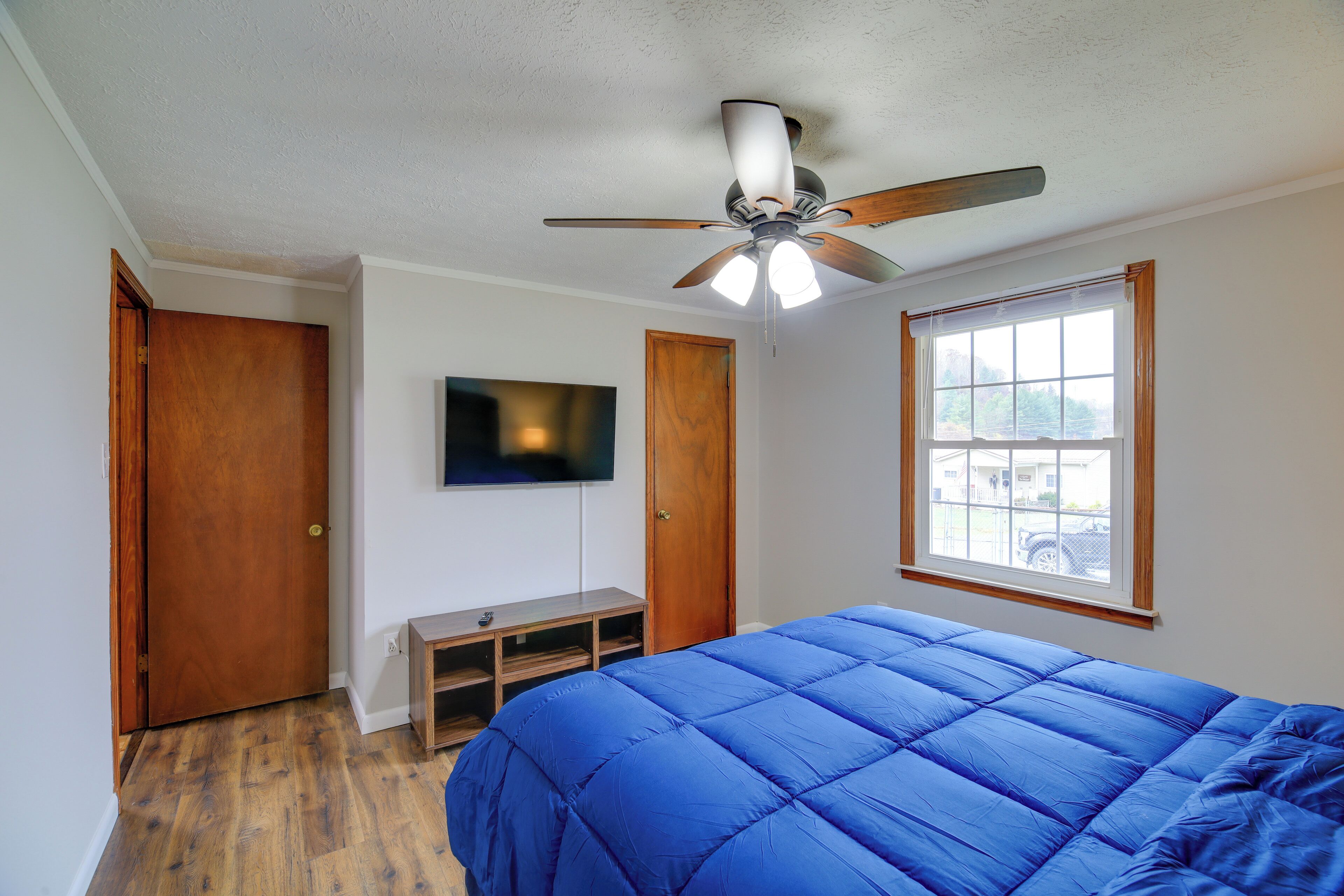 Une chambre avec un ventilateur de plafond, un lit recouvert d’une couverture bleue, un téléviseur fixé au mur et une fenêtre donnant sur l’extérieur.