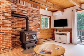A cozy room with a brick fireplace, a television mounted on the wall, and a wooden table with books. - Bartlett Vacation Rental w/ Wraparound Deck! (Bartlett)