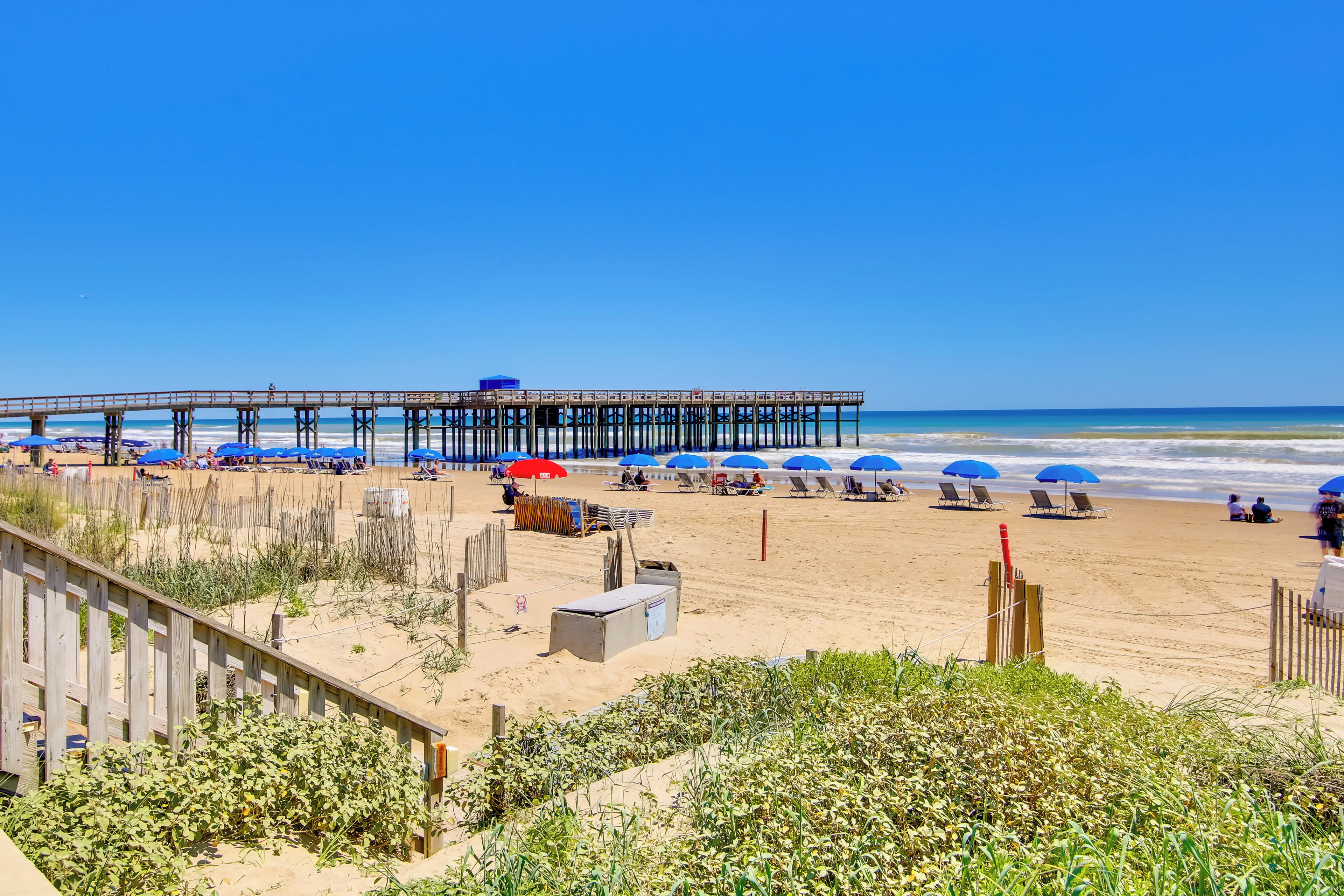 A beach with a wooden pier, beach umbrellas, and lounge chairs.