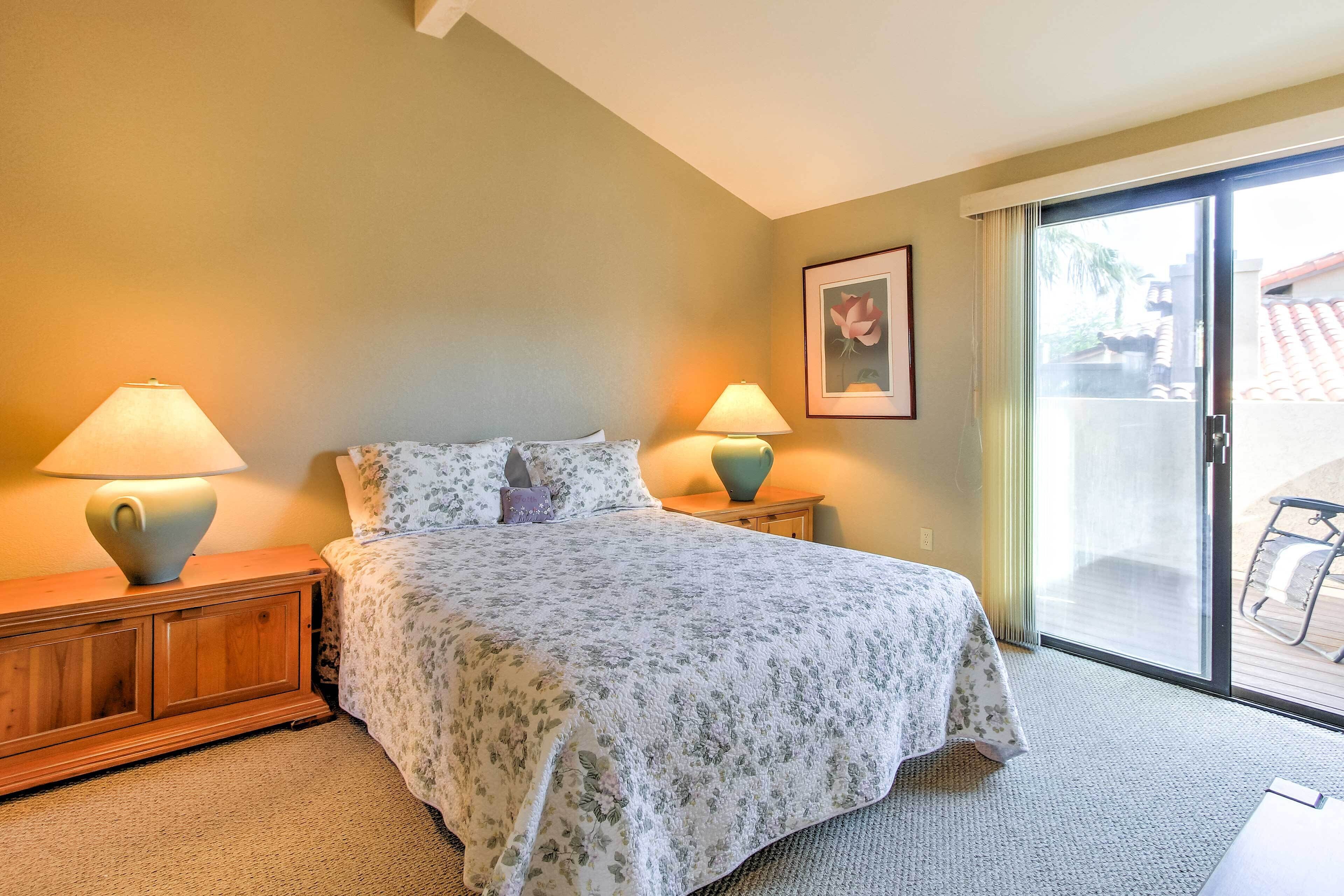 A bedroom with a bed, two bedside tables with lamps, a framed picture on the wall, and a sliding glass door leading to an outdoor area.