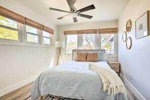A bedroom with a bed, ceiling fan, windows with blinds, bedside table, lamp, and wall decorations.