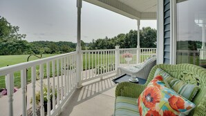 A covered porch with a white railing, wicker furniture, and a view of a green landscape. - Blue Birds Perch: Stunning Home on Private Hilltop (Shipshewana)