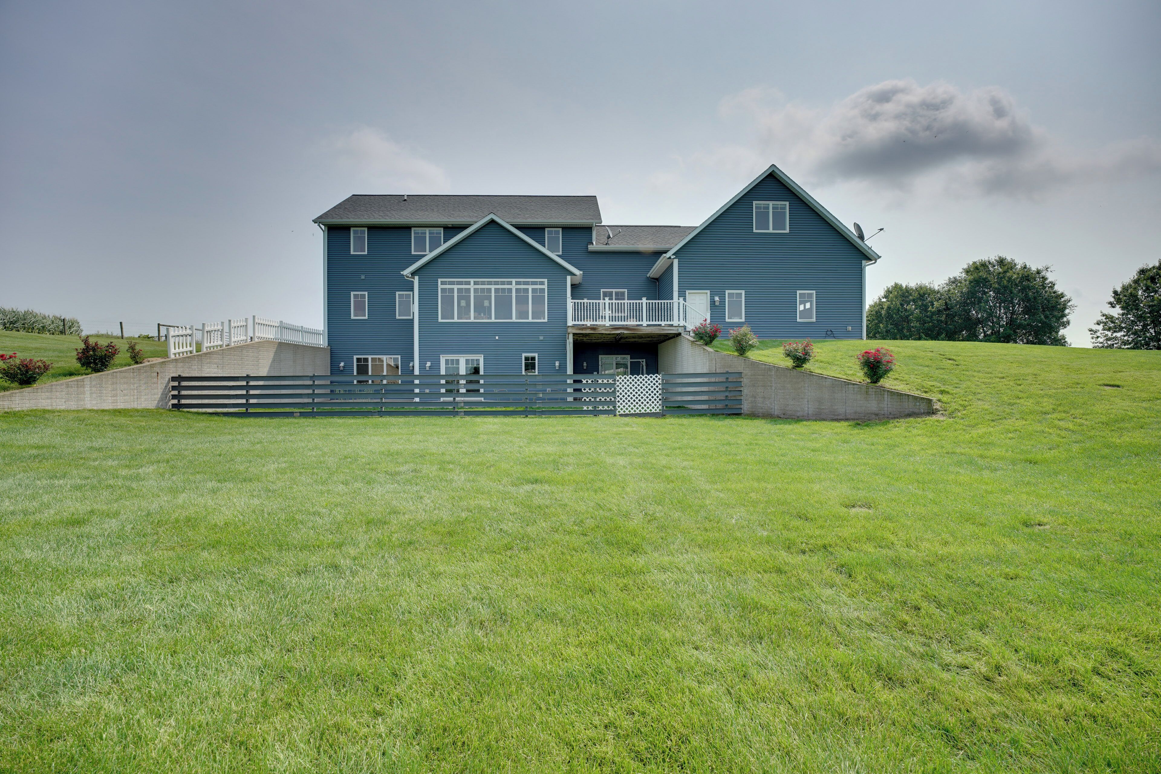 A large blue house with a front porch and a small deck, surrounded by a well-maintained lawn and a stone wall.