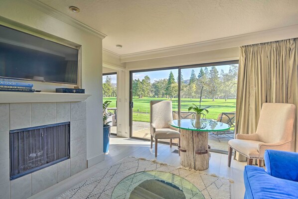 A spacious living room with a fireplace, a television, and a large window offering a view of a golf course. - Bright & Airy Napa Condo W/patio on Golf Course! (Napa)