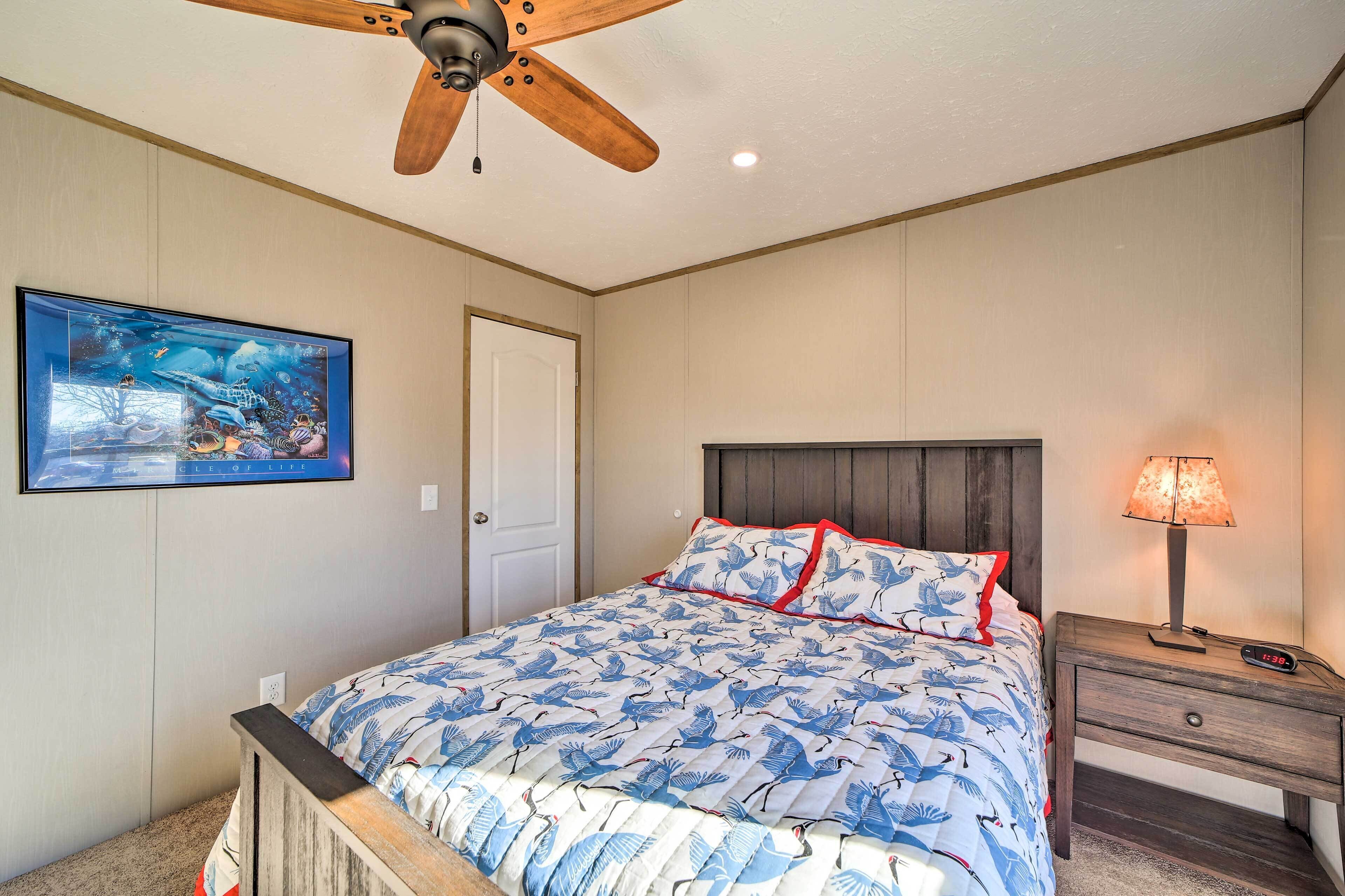 A bedroom with a bed, a ceiling fan, a nightstand with a lamp, and a wall-mounted painting.