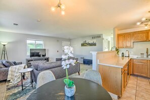 A modern living room with a sofa, a small round table, and a potted orchid. There is a kitchen area with wooden cabinets and a countertop.