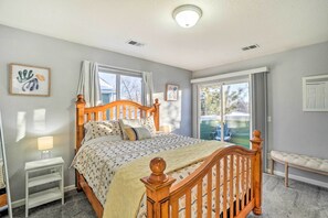 Quarto com cama de madeira, mesa de cabeceira, um banquinho e janela com cortinas.