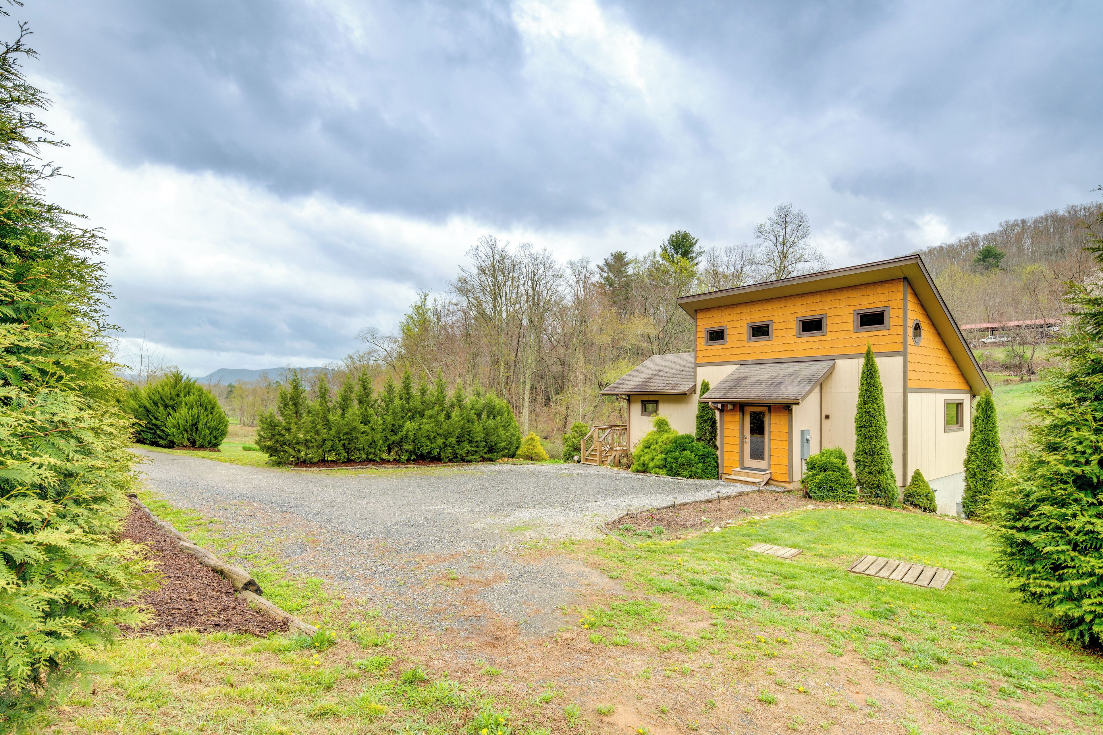 Una casa con camino de grava, un jardín delantero con terraza de madera y un cerro boscoso al fondo.