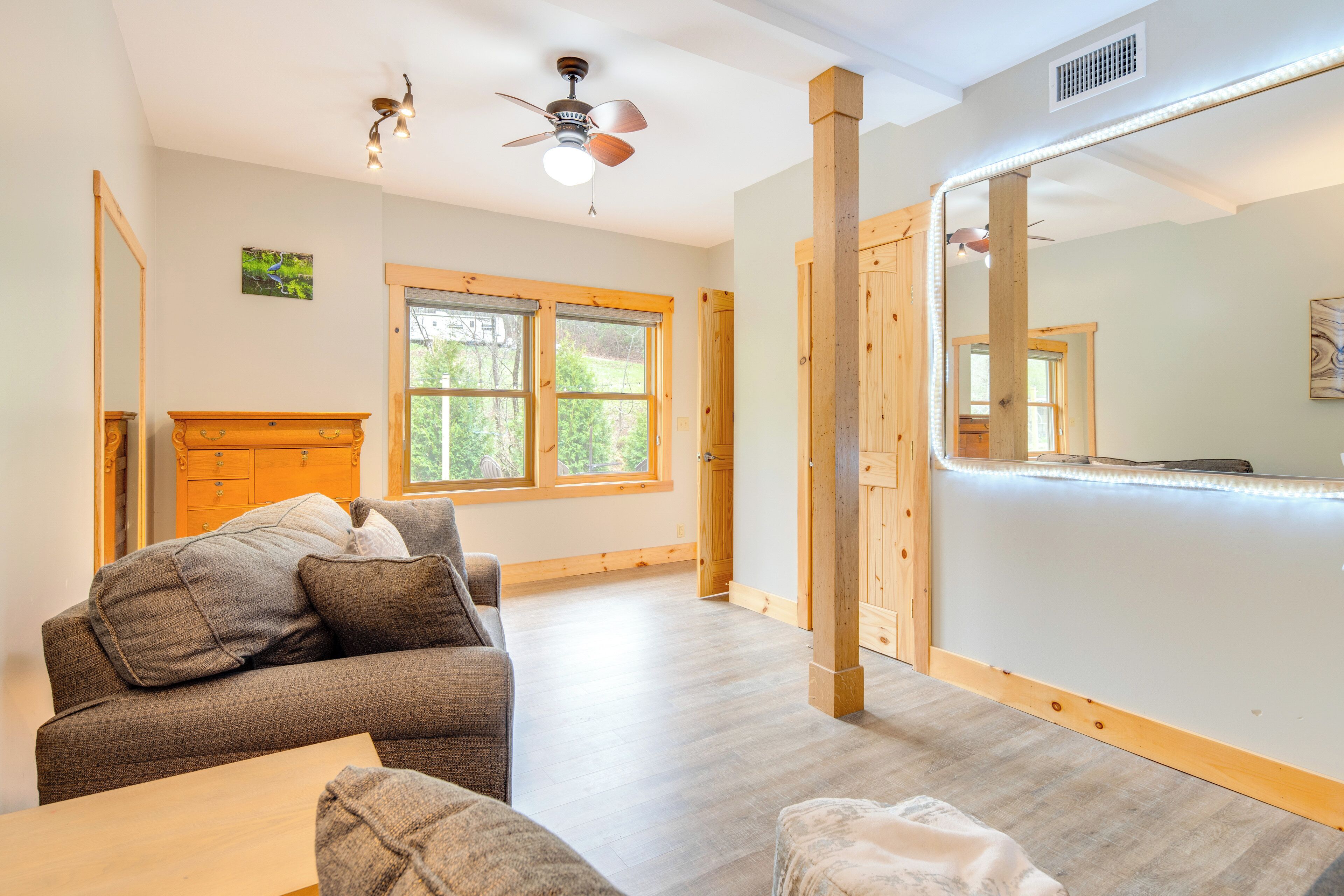 Una habitación amplia con un espejo grande, vigas de madera, ventilador de techo y ventana con vista a la vegetación.