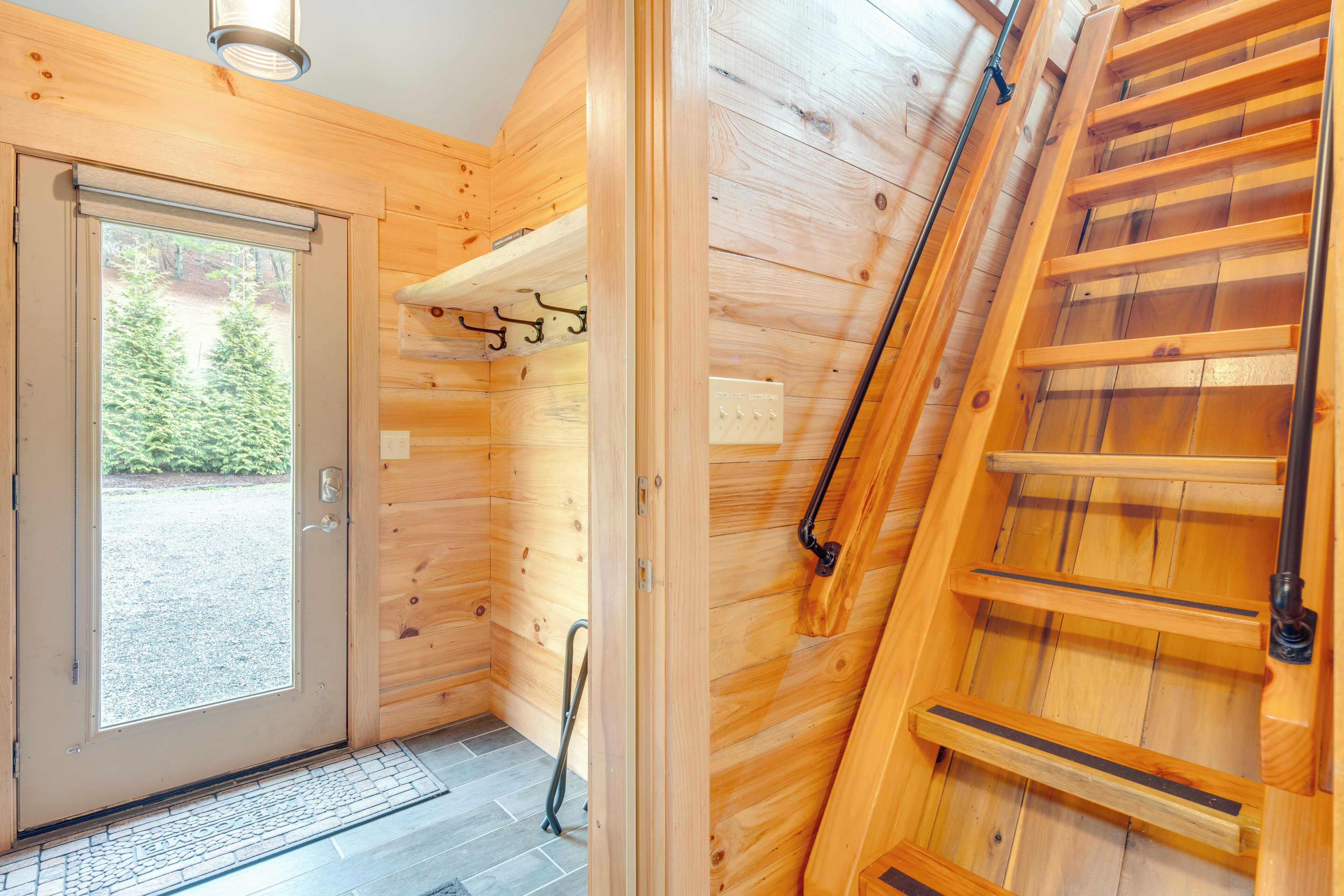 A wooden cabin interior with a door leading outside, a small shelf with hooks, and a wooden staircase.