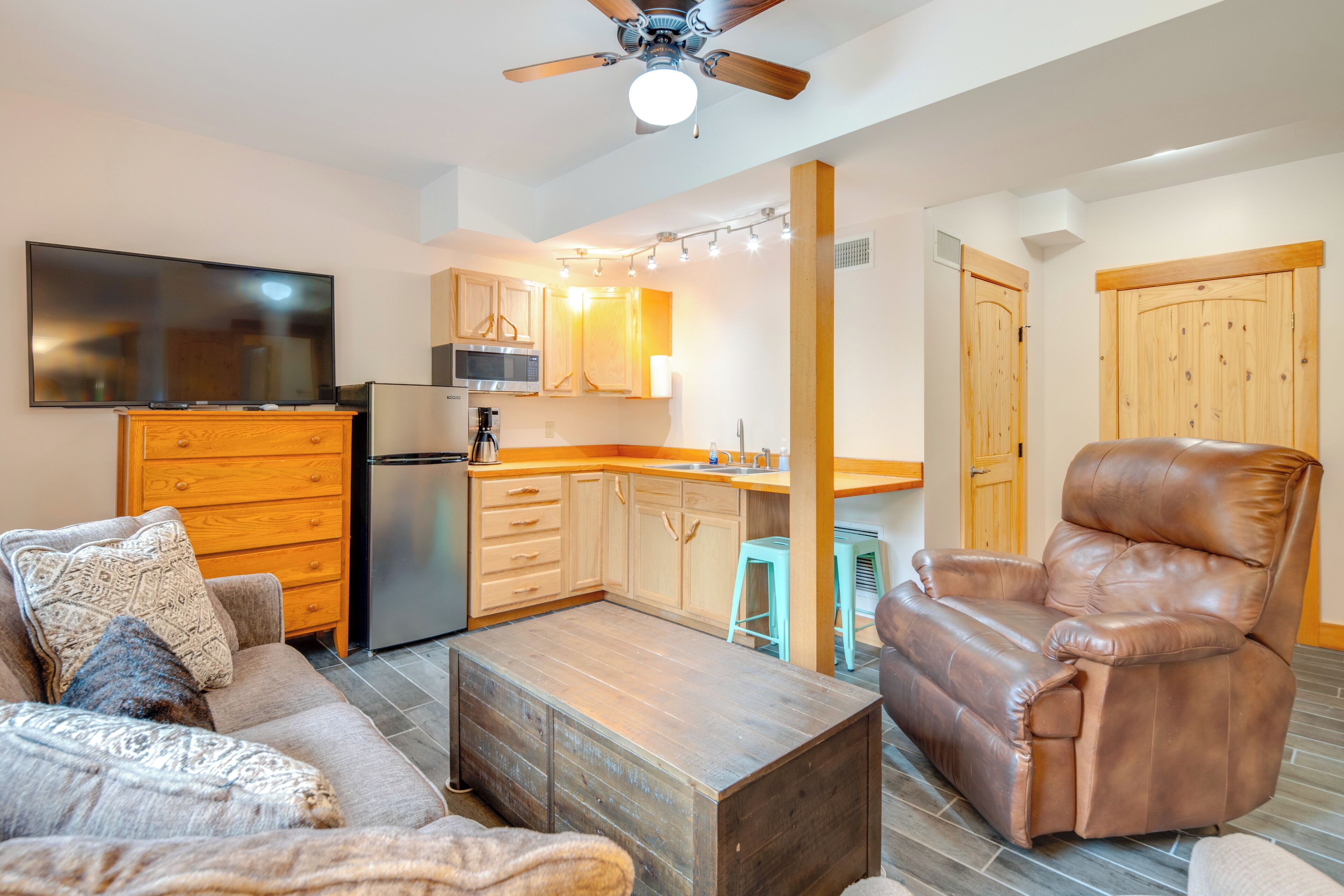 A modern living room with a sofa, coffee table, and leather recliner. A kitchen with wooden cabinets and a microwave is visible in the background.