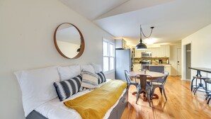 A bedroom with a bed, bedside table, and a large window. There is a kitchen area with cabinets, a microwave, and a dining table with chairs.