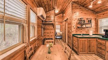 Interior de una cabaña de madera con una mesa de comedor, sillas, zona de cocina y una ventana con persianas.