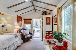 A cozy bedroom with a bed, a chair, a table with a red mug, and a window with curtains. - Charming Tiny Home w/ Deck & Pond Access! (Grass Valley)