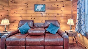 A wooden-paneled room with a leather sofa, two lamps, a bear-themed wall decoration, and patterned curtains. - Charming Jamestown Cabin w/ Large Backyard! (Jamestown)