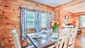 A wooden cabin interior with a dining table set for four, flanked by two windows with patterned curtains. - Charming Jamestown Cabin w/ Large Backyard! (Jamestown)
