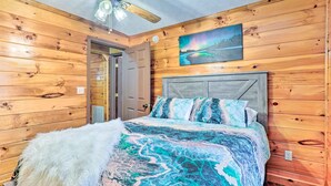 A wooden-paneled room with a bed featuring a patterned bedspread and pillows, a ceiling fan, and a painting on the wall. - Charming Jamestown Cabin w/ Large Backyard! (Jamestown)