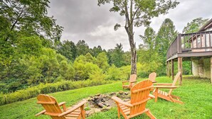 A deck with four wooden Adirondack chairs facing a fire pit in a lush green garden. - Charming Jamestown Cabin w/ Large Backyard! (Jamestown)