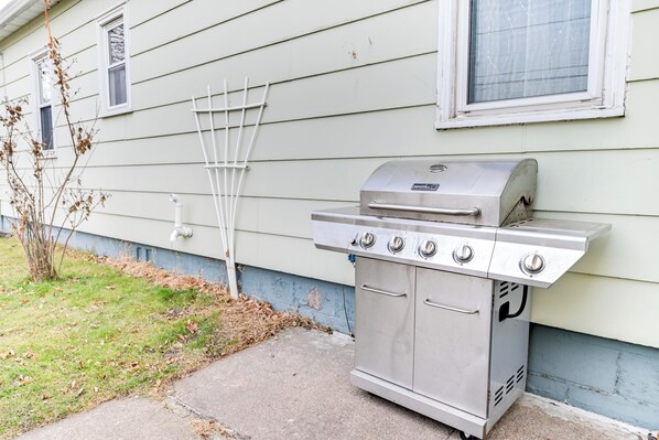 En grill i rostfritt stål med fyra brännare och en sidobrännare, placerad på en betongterrass framför ett hus med vit träpanel och ett fönster.