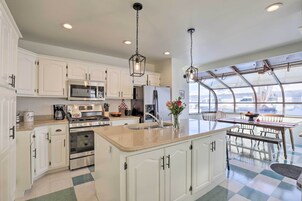 Una cocina con gabinetes blancos, una isla central, electrodomésticos de acero inoxidable y un comedor con vistas a través de un cerramiento de vidrio.