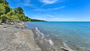 A pebble beach with clear water, trees, and a distant shoreline. - Charming Lake Erie Getaway: Walk to Beach! (North East)