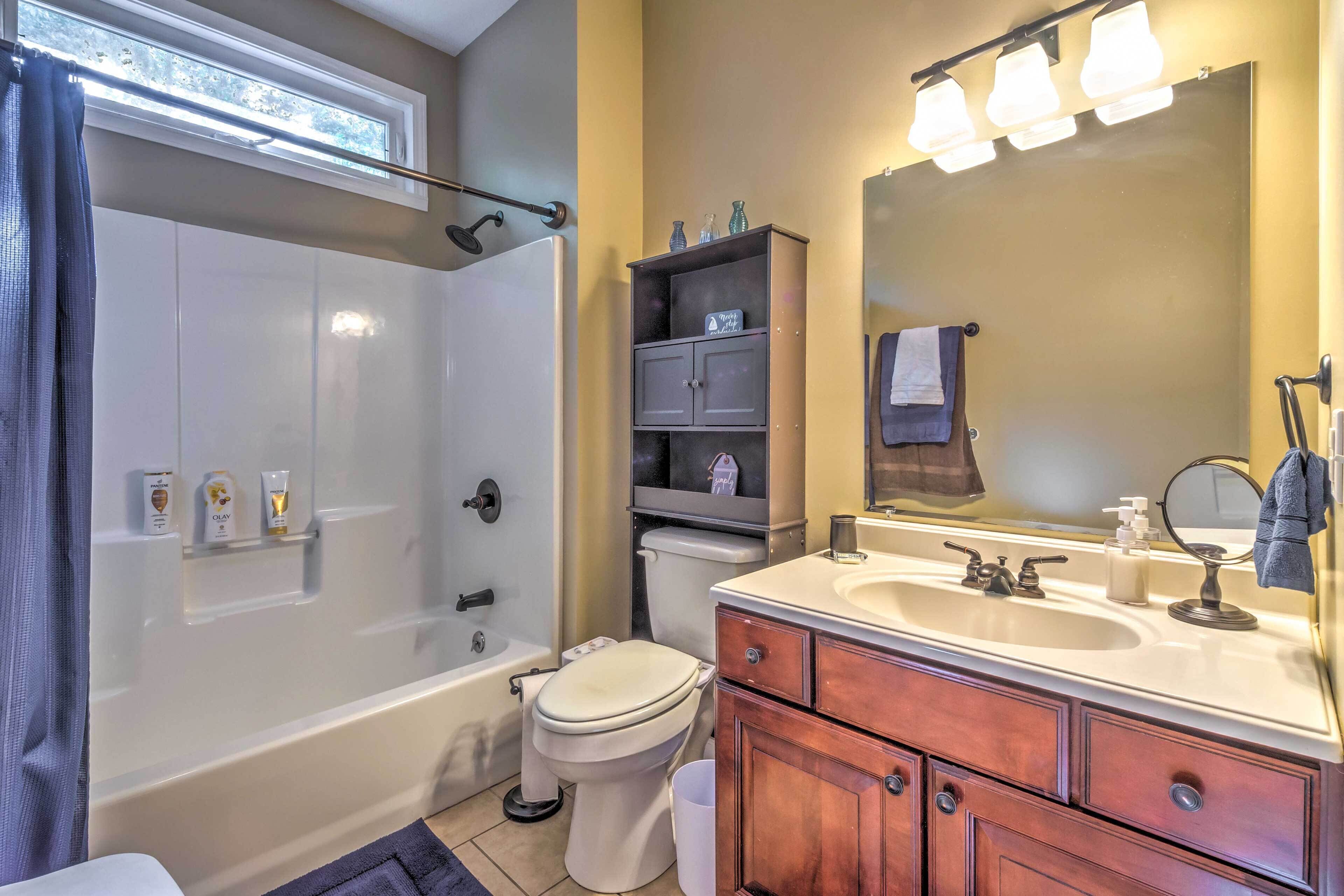 A bathroom with a bathtub, toilet, vanity with drawers, a mirror, and shelves with toiletries.
