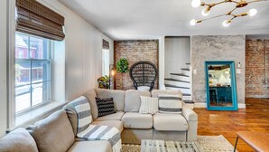 A modern living room with a sectional sofa, a black woven chair, a staircase, and a kitchen visible through a mirrored door. - Chic Troy Home Near Downtown, 9 Mi to Albany! (Troy)
