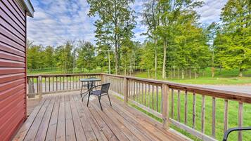 A wooden deck with a table and chairs, surrounded by a wooden railing, overlooking a grassy area with trees.