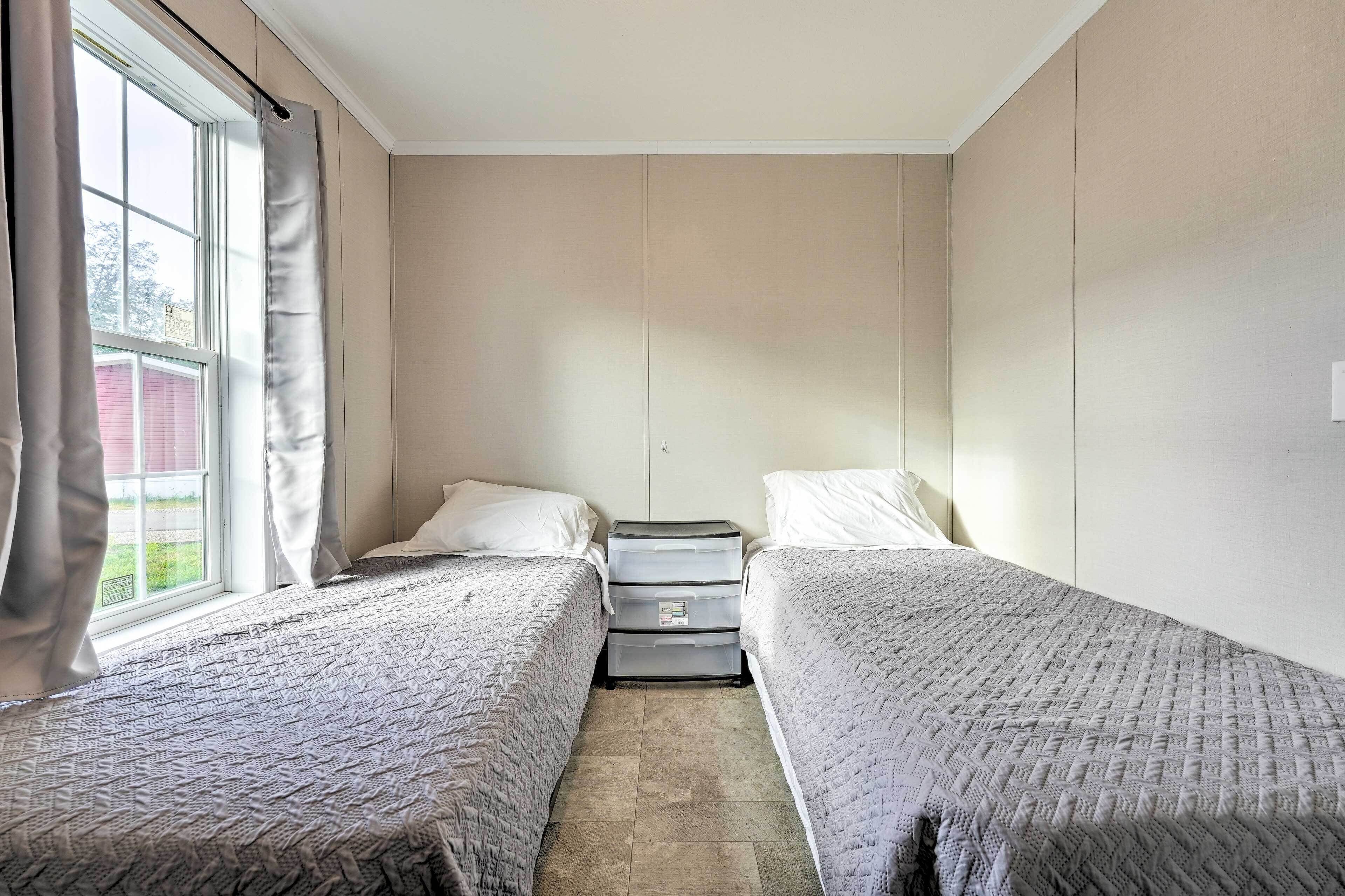 A room with two single beds, a nightstand with drawers, a window with curtains, and a view of a red building outside.