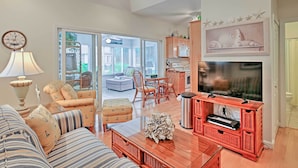 A living room with a sofa, armchair, and ottoman, a television on a wooden stand, and a dining area with a table and chairs.