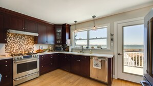A modern kitchen with dark wood cabinets, a large stainless steel oven, a microwave, and a view of the ocean through a sliding glass door. - Welcoming Cottage w/ Outdoor Pool & Water Views! (White Stone)