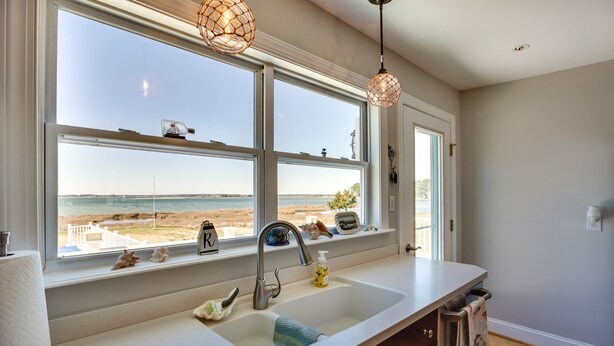 A kitchen with a large window overlooking a beach, a sink, and a countertop with various items. - Welcoming Cottage w/ Outdoor Pool & Water Views! (White Stone)