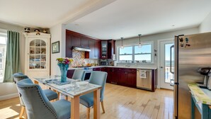 A modern kitchen with wooden cabinets, a stainless steel refrigerator, and a dining area with a glass table and blue chairs. - Welcoming Cottage w/ Outdoor Pool & Water Views! (White Stone)