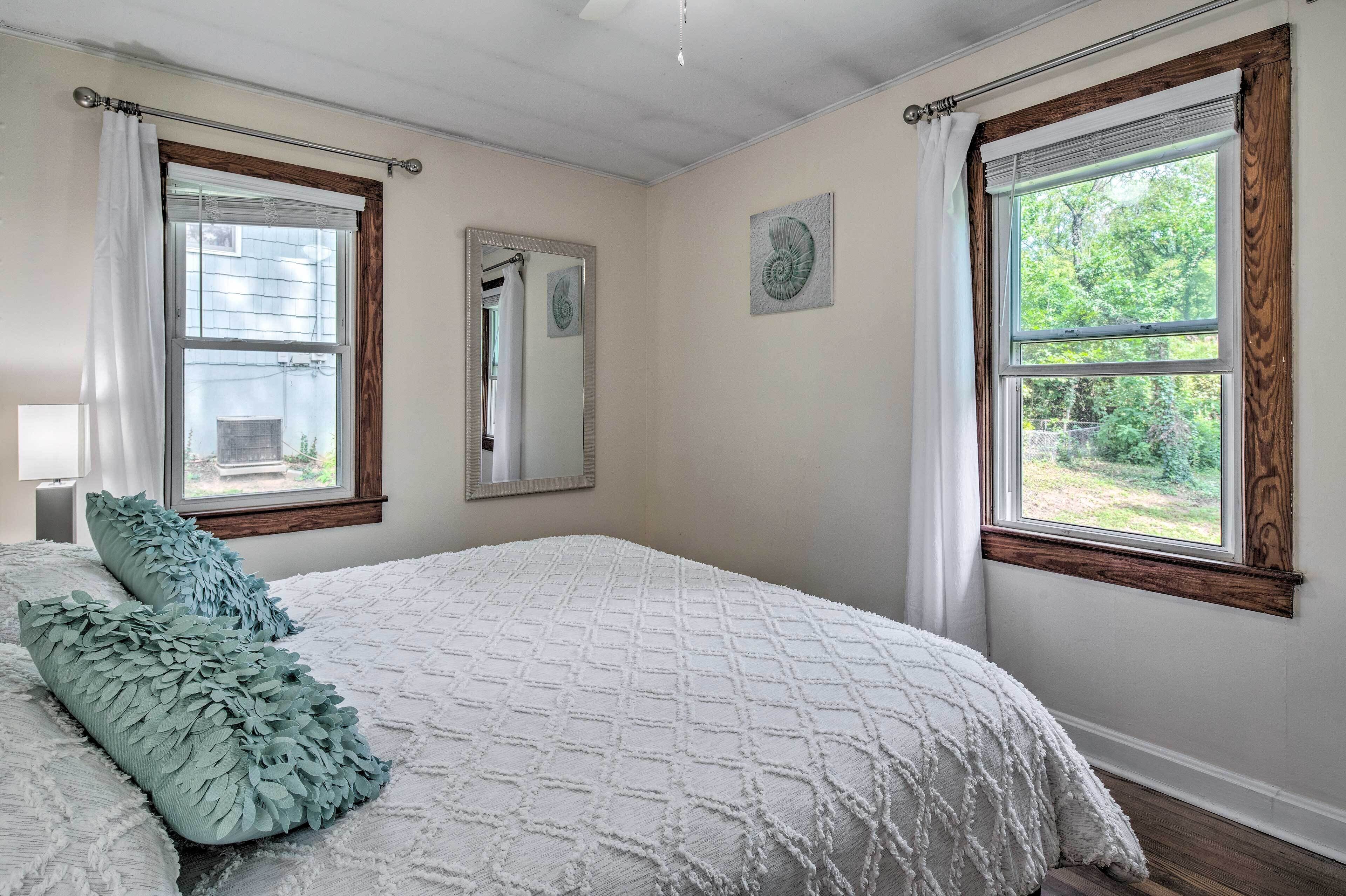 A bedroom with a large bed, two windows with wooden frames, a mirror, and a view of greenery outside.