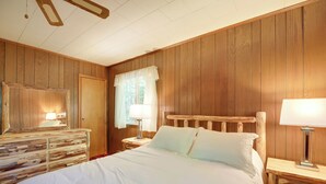 A wooden-paneled bedroom with a bed, a dresser, and a nightstand with a lamp. - Cozy Adirondacks Cabin: Walk to Lake Eaton! (Jay)