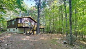 A cabin with a wooden deck and a staircase leading to a second level, surrounded by a forested area. - Cozy Adirondacks Cabin: Walk to Lake Eaton! (Jay)