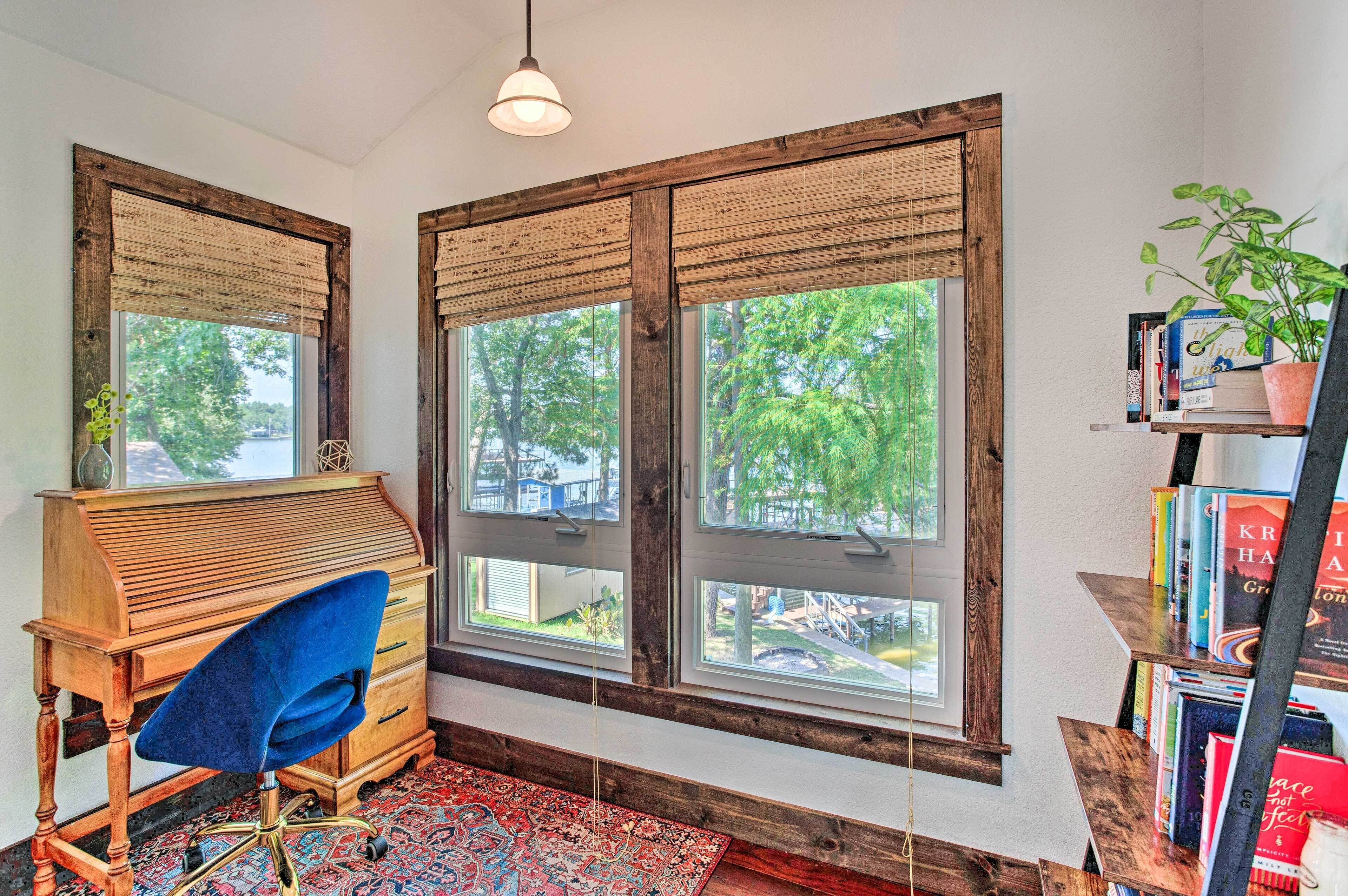 A room with large windows, a wooden desk, a blue chair, a bookshelf, and a patterned rug.