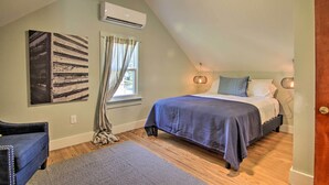 A bedroom with a sloped ceiling, a bed with a blue bedspread, a chair, a rug, and a window with curtains. - 'cora's Cottage' Near Chesapeake Bay Access! (Harborton)