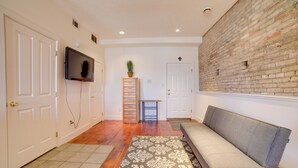 Ein Flur mit grauem Sofa, einem gemusterten Teppich, Holzdielen, einem an der Wand montierten Fernseher und einer Ziegelwand.