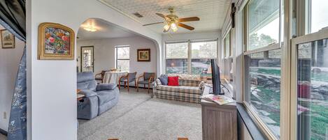 A spacious living room with a ceiling fan, large windows, a sofa, a coffee table, and a desk.