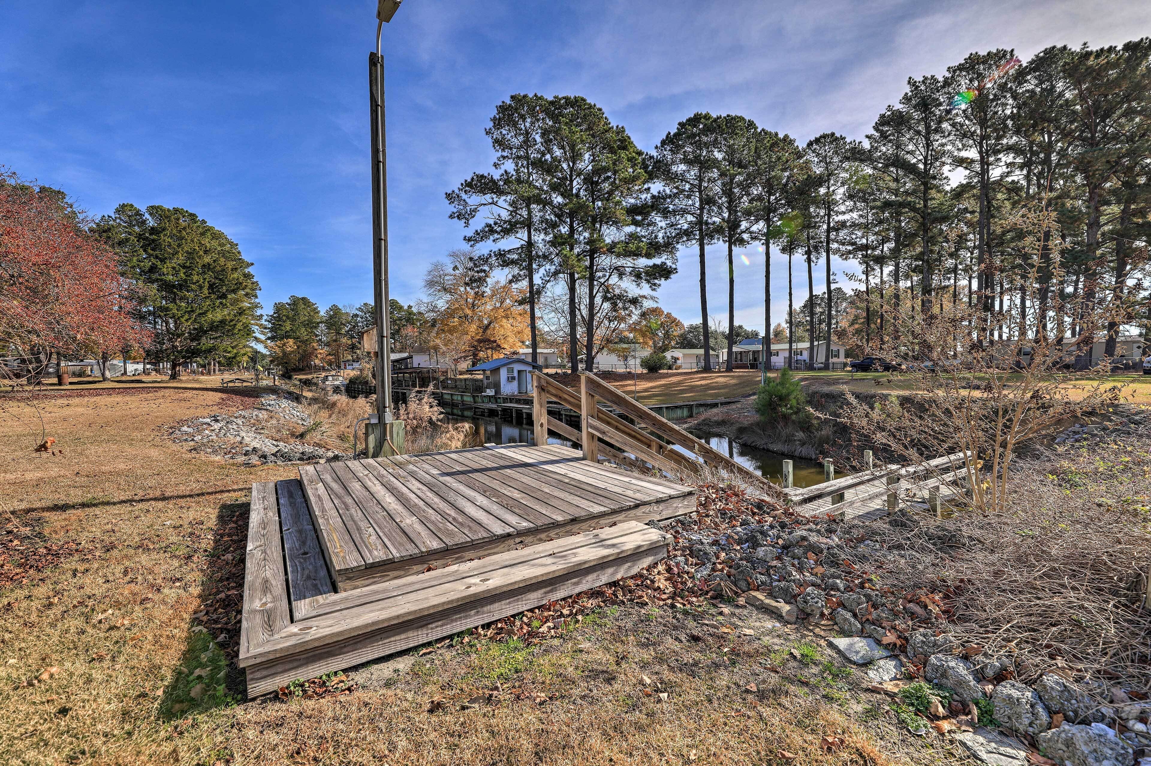 A wooden deck leading to a small bridge over a water feature, surrounded by trees and a grassy area.
