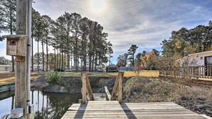 Un pont en bois menant à un quai surplombant une étendue d’eau calme, entouré de grands arbres et sous un ciel dégagé.